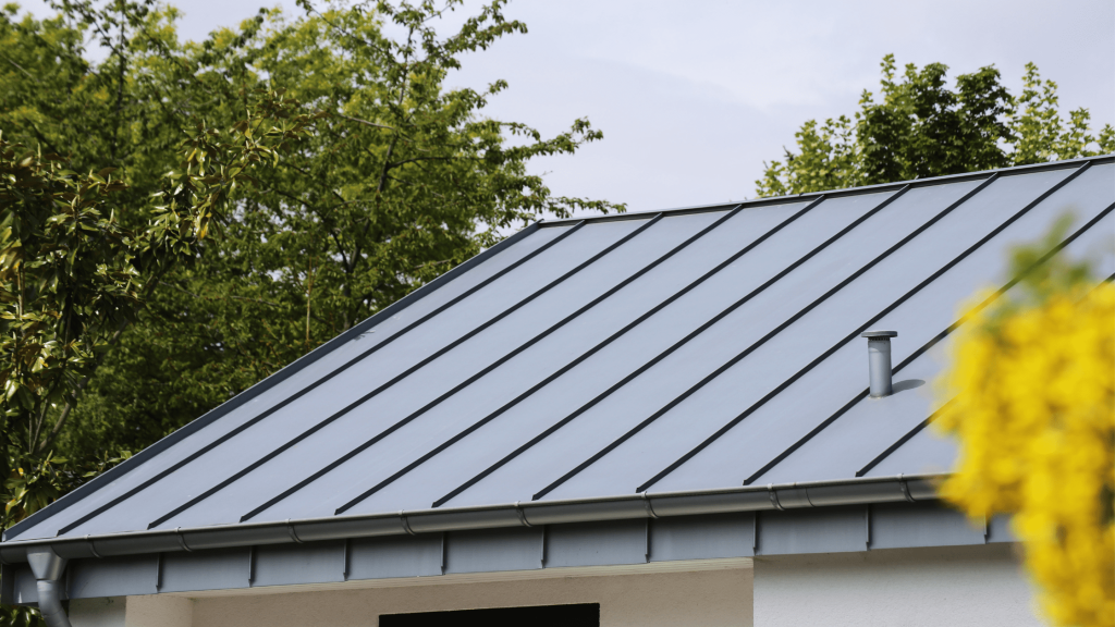 A close-up view of a standing seam metal roof on a house, with a background of green trees and a clear sky. The roof features clean, parallel lines and a sleek, modern appearance. A small ventilation pipe is visible on the right side of the roof.