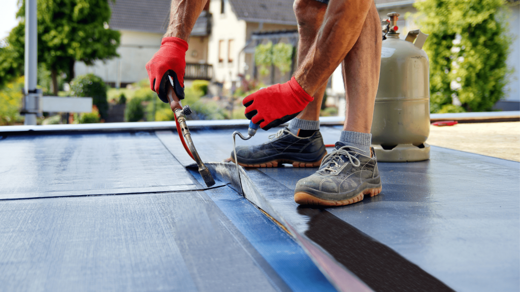 Person working on a flat roof, wearing red gloves and using a blowtorch to seal roofing material, with a gas tank visible in the background. The setting appears to be a residential area with greenery and houses in the background.
